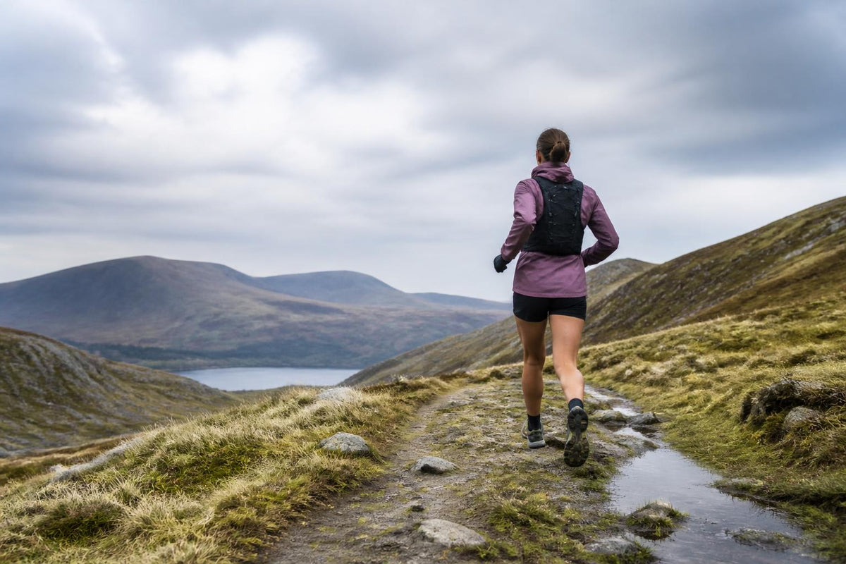 A runner wearing a lightweight trail vest and weather-resistant gear stands on a rocky mountain path
